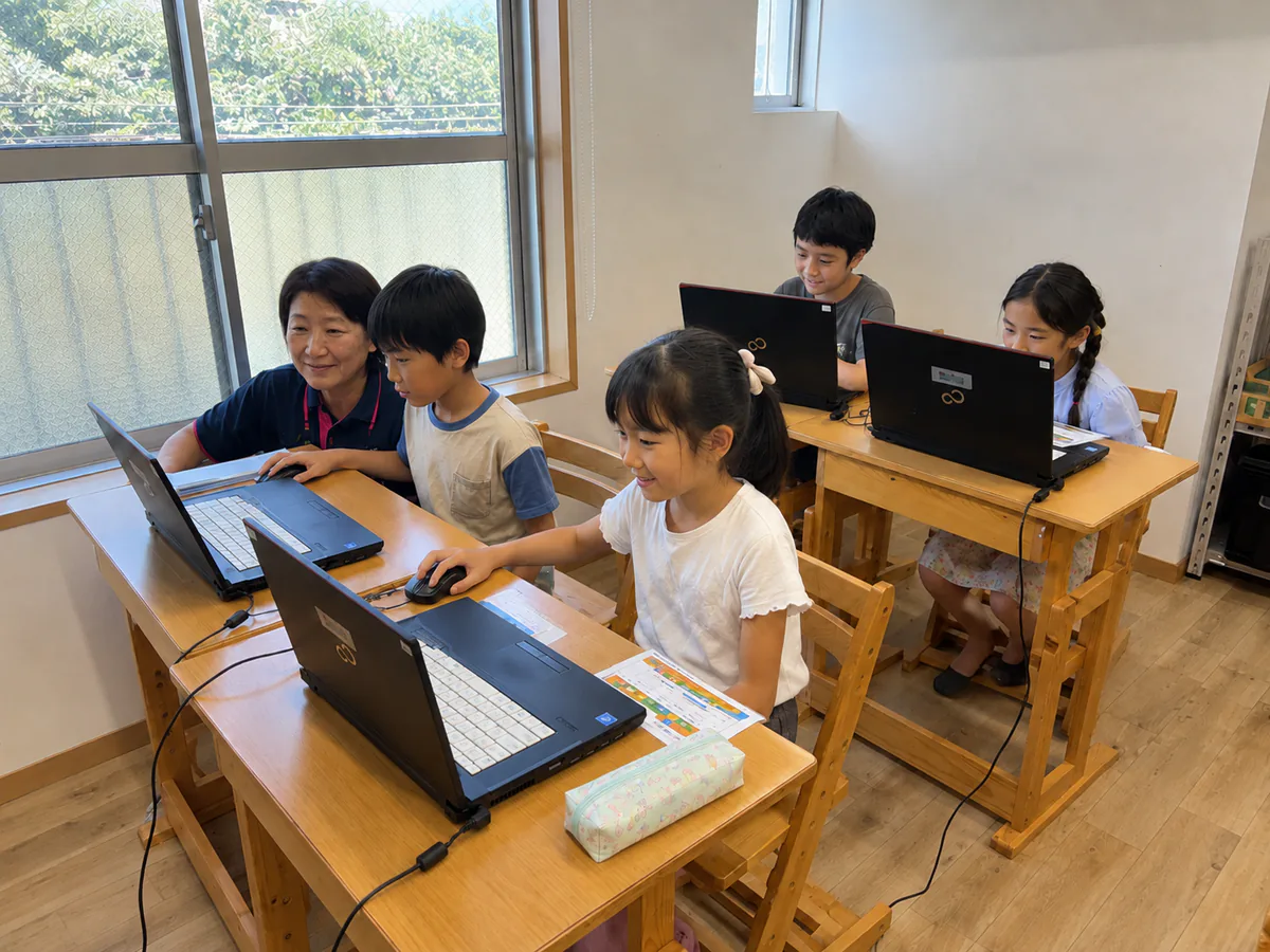 学童の教室で講師と子どもたちがノートパソコンを使ってプログラミングに取り組む様子