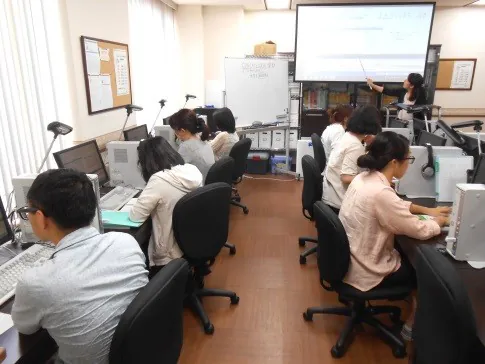 教室でデスクトップパソコンに向かって受講する訓練生と、スクリーンの前で解説する講師
